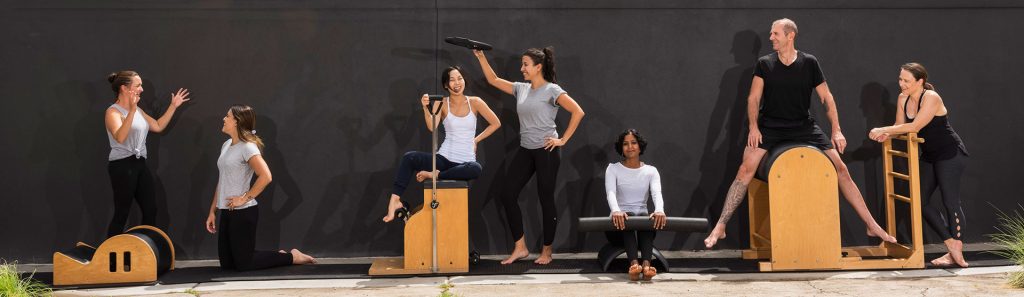 Movement Principle pilates instructors goofing around on pilates equipment outside against dark grey wall