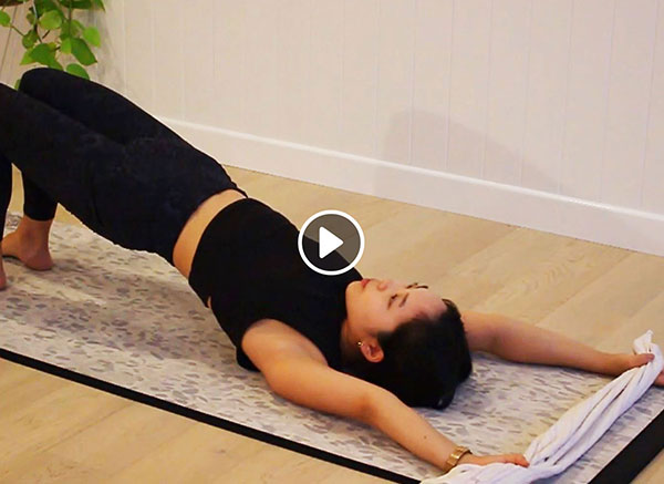 Pilates instructor doing pelvic lift on floor holding towel above head with play button icon