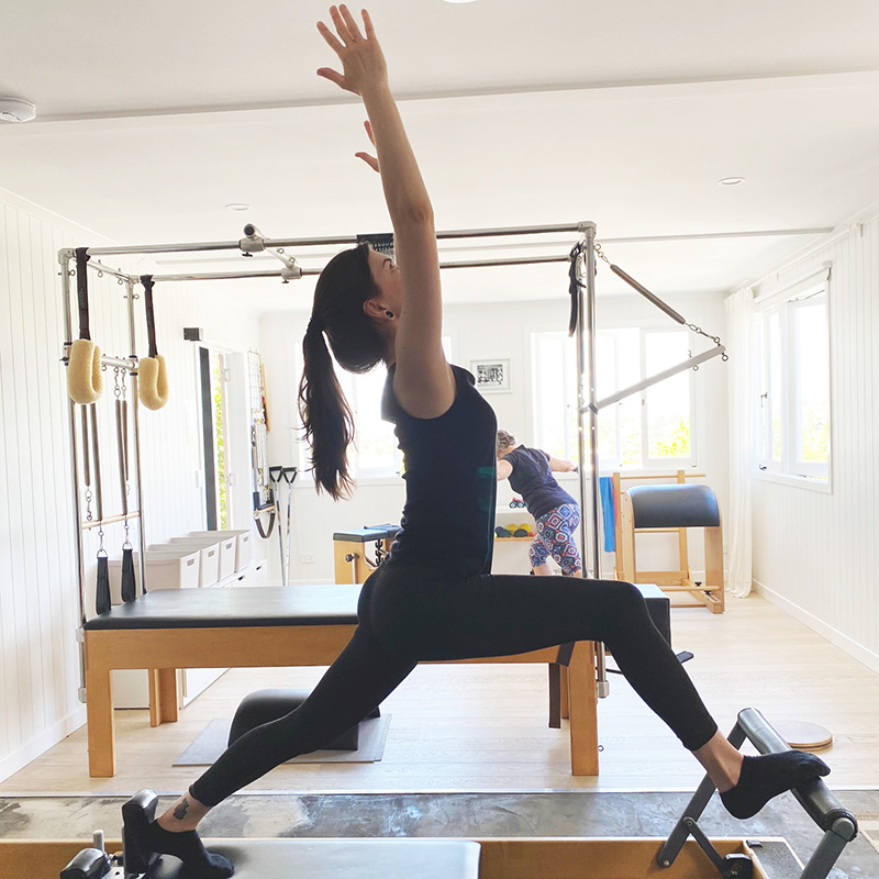 young woman wearing black gym clothes in pilates studio lunging on reformer machine with arms stretched up