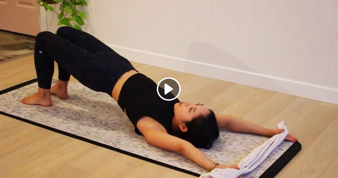Pilates instructor doing pelvic lift on floor holding towel above head with play button icon